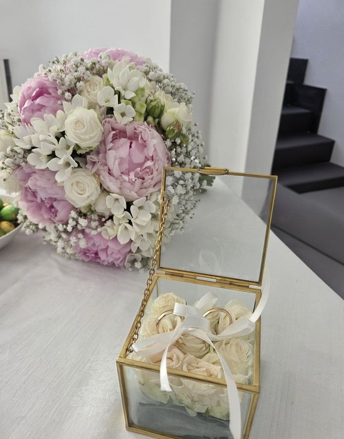 Portafedi in vetro e ottone con le fedi su un letto di roselline bianche, accanto a un bouquet di peonie rosa, rose e fiori bianchi