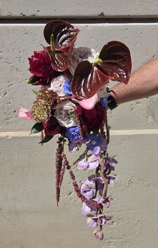 Bouquet a cascata con anthurium bordeaux, rose, peonia, delphinium e orchidee lilla su fondo muro in cemento