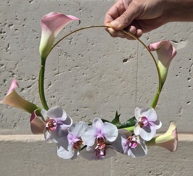 Bouquet moderno ad anello dorato con calle rosa e orchidee phalaenopsis bianche e lilla, tenuto in mano davanti a un muro di pietra