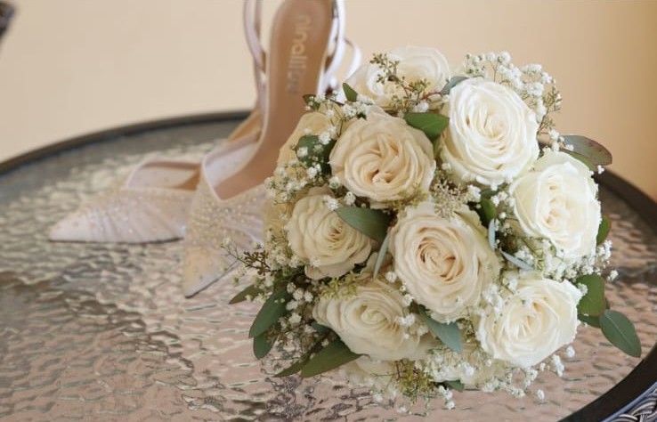 Bouquet tondo di rose bianche, gypsophila ed eucalipto appoggiato su un vassoio accanto alle scarpe da sposa