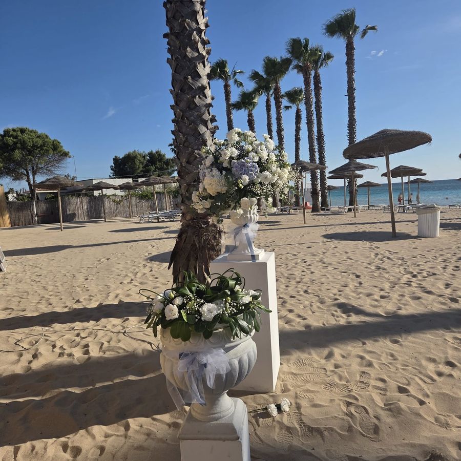 Allestimento di cerimonia in spiaggia con vasi classici e composizioni di ortensie azzurre, rose e lisianthus bianchi su colonne
