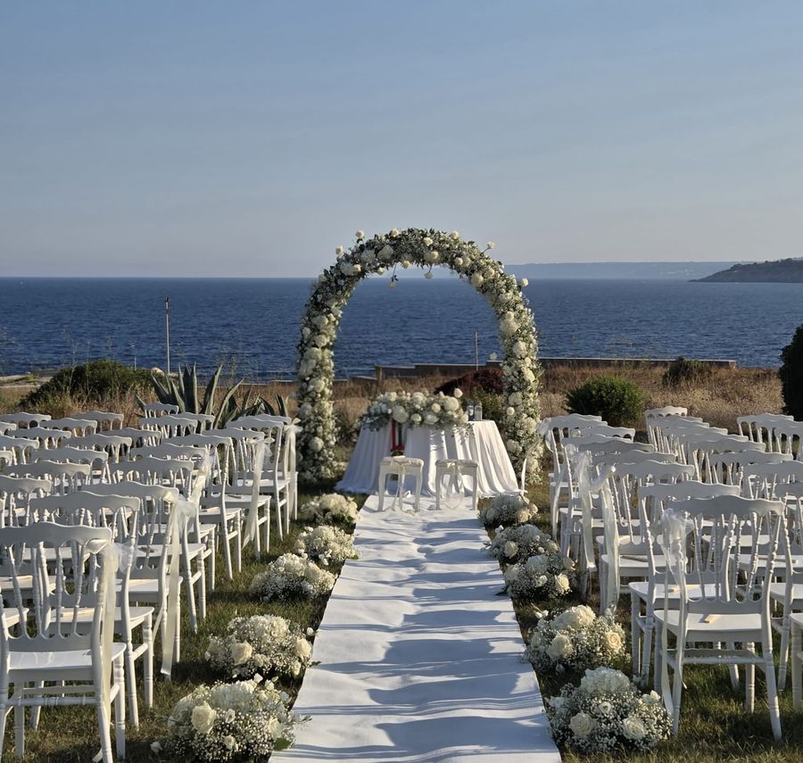 Cerimonia all'aperto sul mare con arco di fiori bianchi, passerella bianca tra file di sedie bianche e composizioni di rose e ortensie bianche a terra