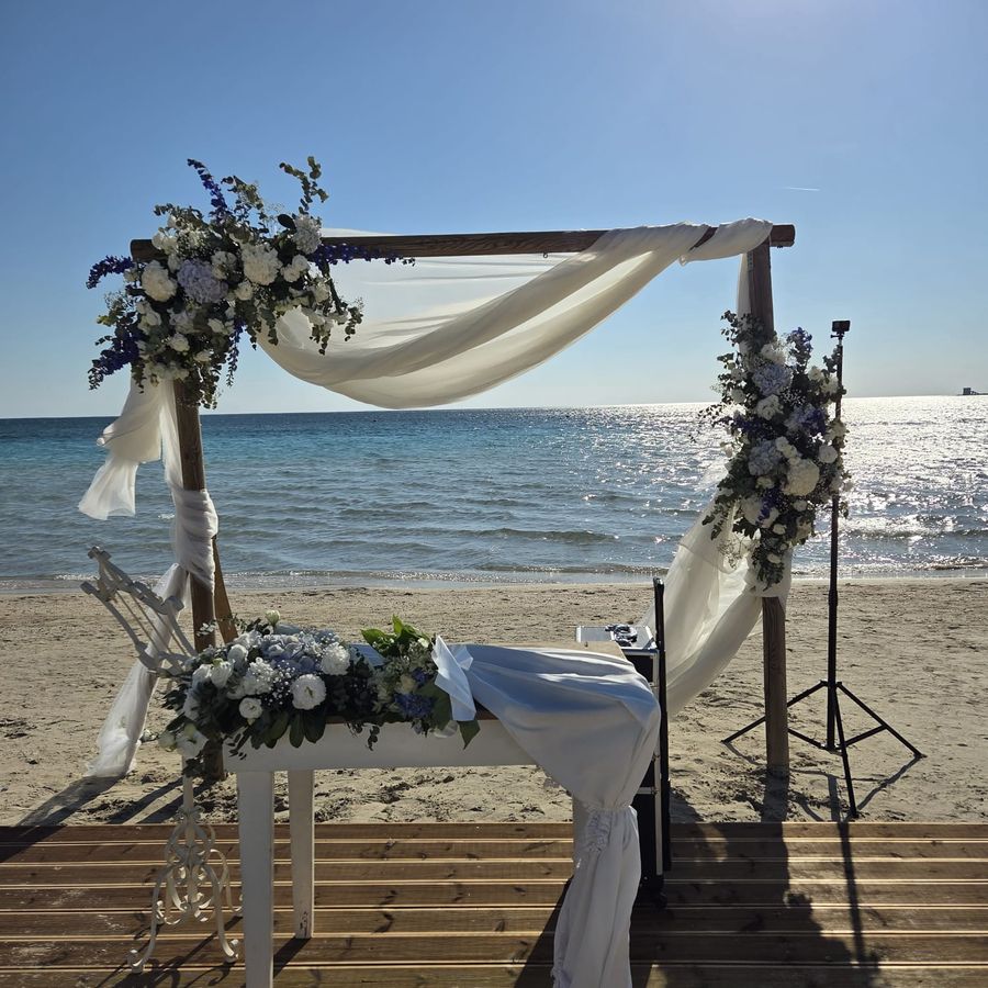 Arco nuziale in legno sulla spiaggia con veli bianchi e composizioni di ortensie, rose e delphinium azzurri, con tavolo della cerimonia decorato di fiori davanti al mare