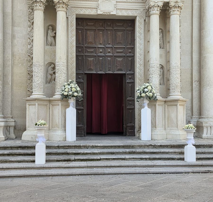 Ingresso di una chiesa barocca leccese con colonnine bianche e composizioni di fiori bianchi ai lati del portone e lungo la scalinata