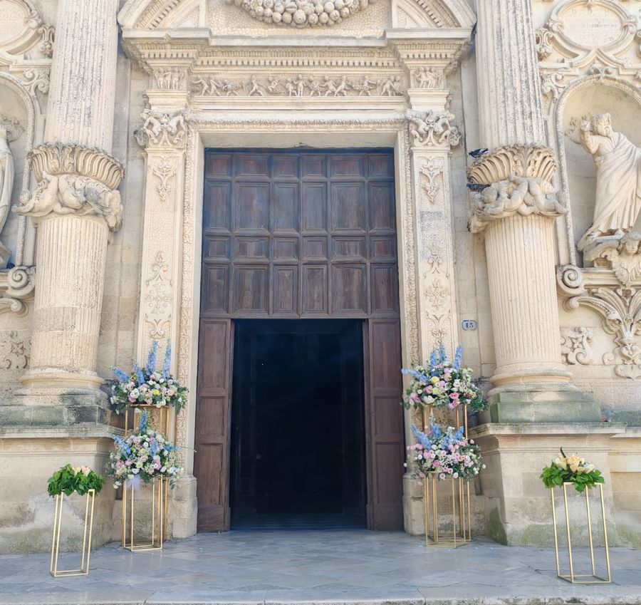 Portale barocco di una chiesa leccese con alzate dorate e composizioni di delphinium azzurro, rose e fiori rosa ai lati dell'ingresso