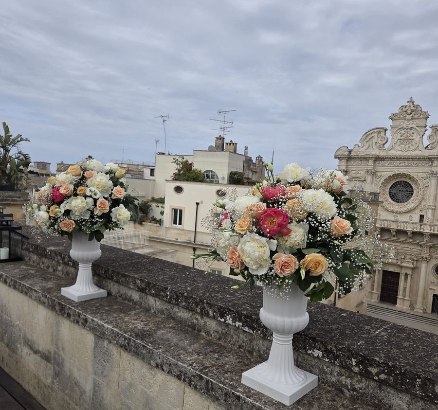 Coppia di composizioni di rose pesca, fucsia e bianche con gypsophila in vasi bianchi a calice su un muretto, con la Basilica di Santa Croce di Lecce sullo sfondo