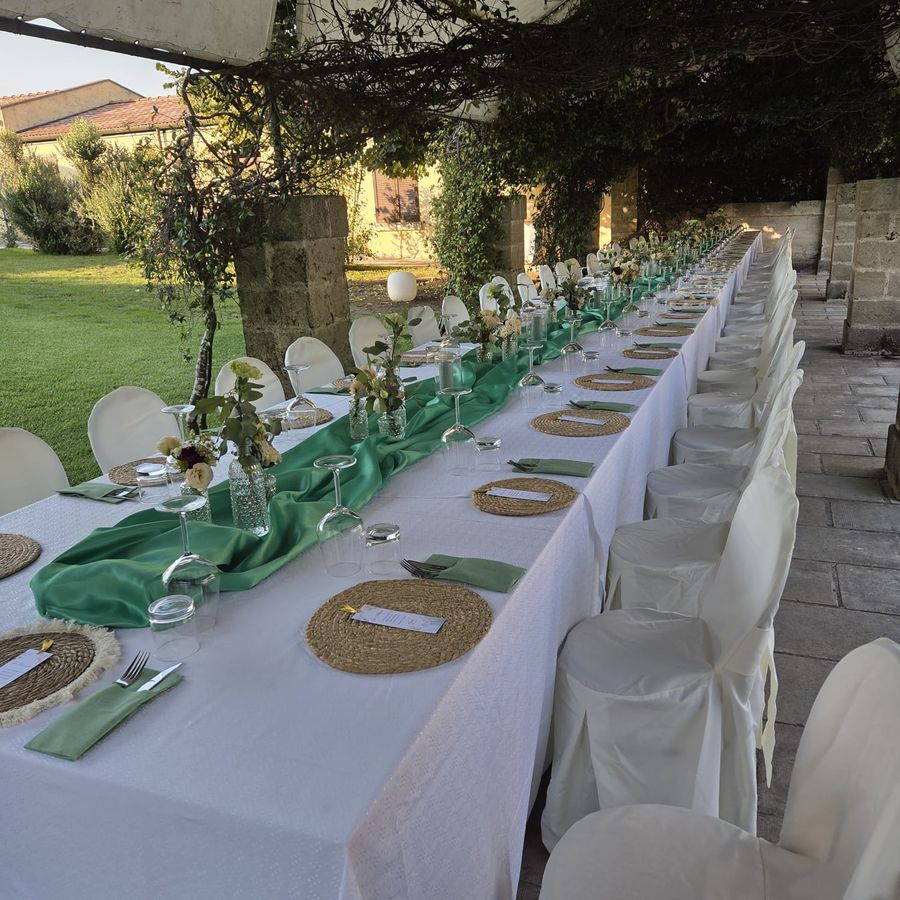Tavolo imperiale allestito sotto un pergolato con runner verde salvia, tovagliato bianco, sottopiatti in rafia e vasetti di fiori bianchi