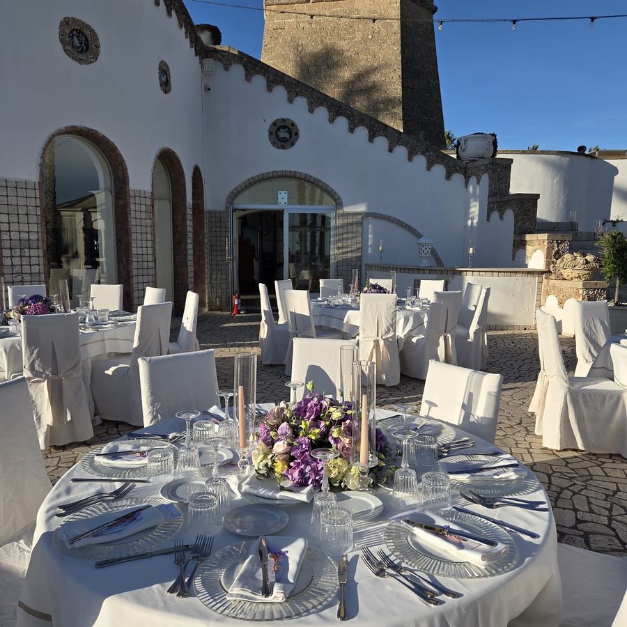 Ricevimento all'aperto in masseria con tavoli rotondi, coprisedie bianchi e centrotavola bassi di ortensie e lisianthus lilla
