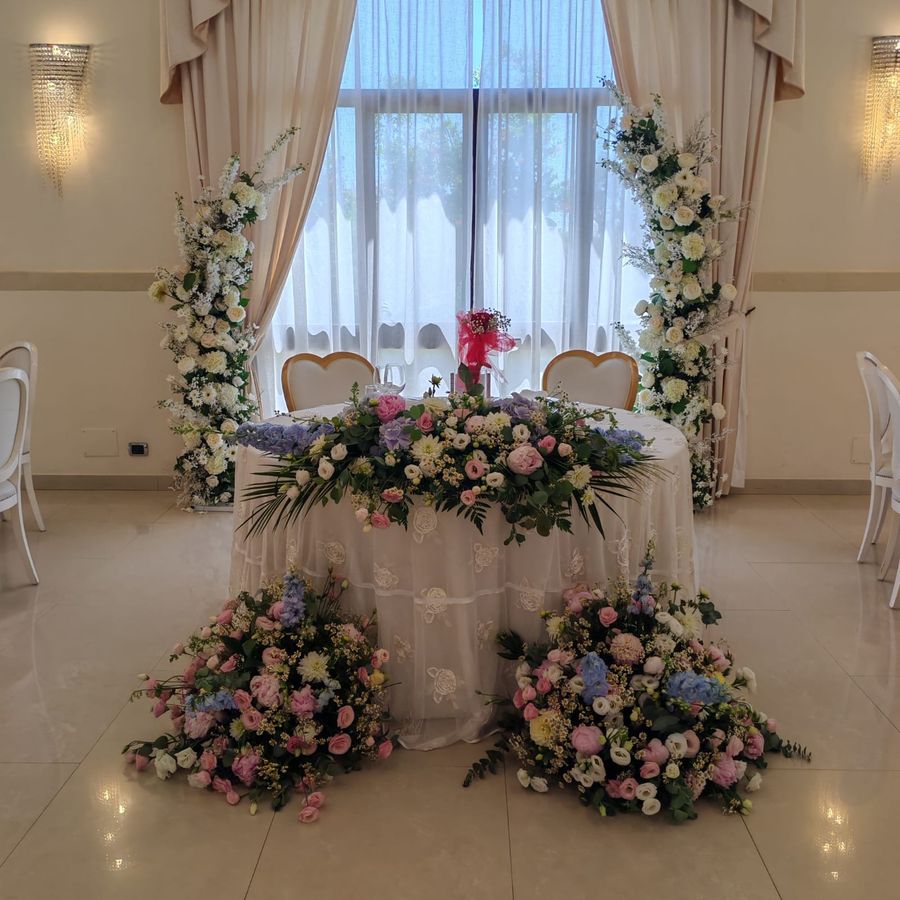 Tavolo degli sposi in sala ricevimento con runner floreale rosa e azzurro, due colonne fiorite laterali e composizioni a terra