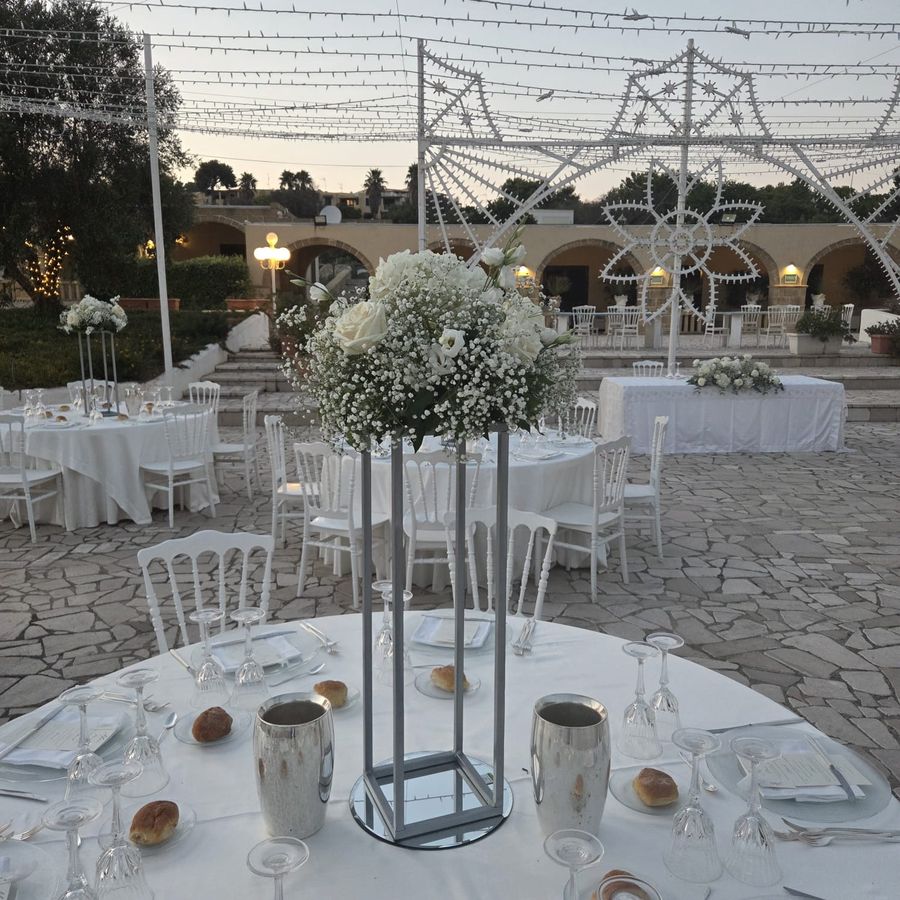 Centrotavola alto di gypsophila e rose bianche su alzata metallica in un ricevimento all'aperto con luminarie e tavolo degli sposi