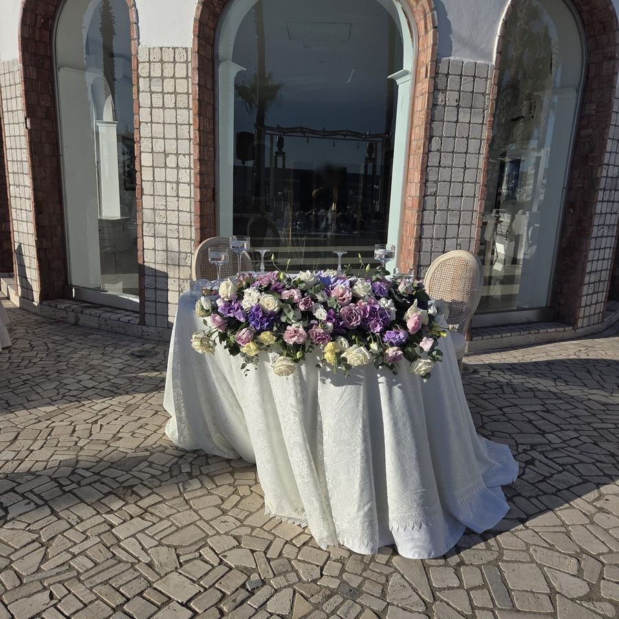 Tavolo degli sposi all'aperto con composizione lunga di rose lilla, viola e bianche su tovaglia bianca ricamata