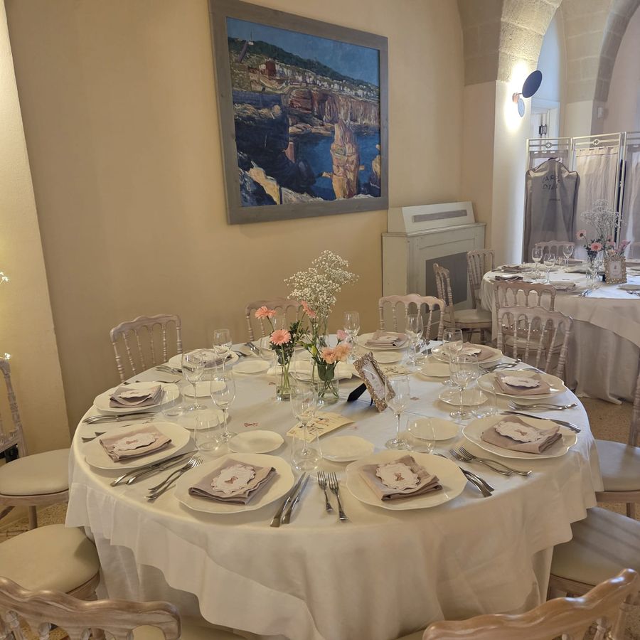 Tavolo rotondo di ricevimento con mise en place completa, tovagliato bianco e piccoli vasi con gerbere rosa e gypsophila